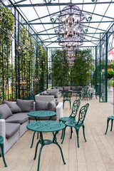 A modern grey sofa with cushions is installed in a glass greenhouse outside. Lush green plants and...