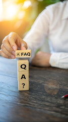 FAQ Block Question: A person arranges wooden blocks spelling out "FAQ" with a question mark, set against a desk and sunlight background. The image conveys questions and answers