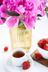A cup of tea sits on a saucer next to a bowl of fresh strawberries. Pink peonies are placed nearby