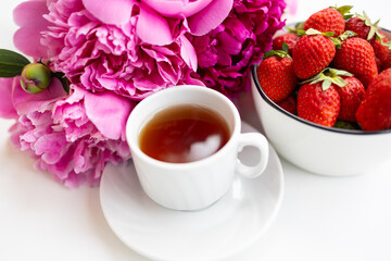 A cup of tea sits on a saucer next to a bowl of fresh strawberries. Pink peonies are placed nearby