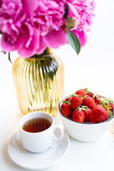 A cup of tea sits on a saucer next to a bowl of fresh strawberries. Pink peonies are placed nearby