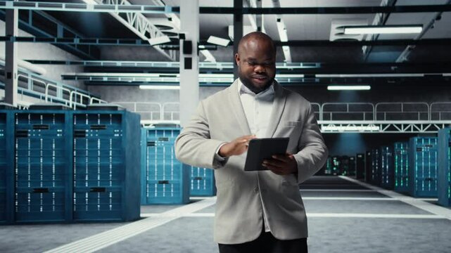 Smiling admin using tablet in in data center maintaining infrastructure for workloads. Joyful IT professional in server farm utilizing high tech server clusters for parallel processing, camera A
