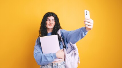 Brunette student girl taking selfie holding notebook on yellow background
