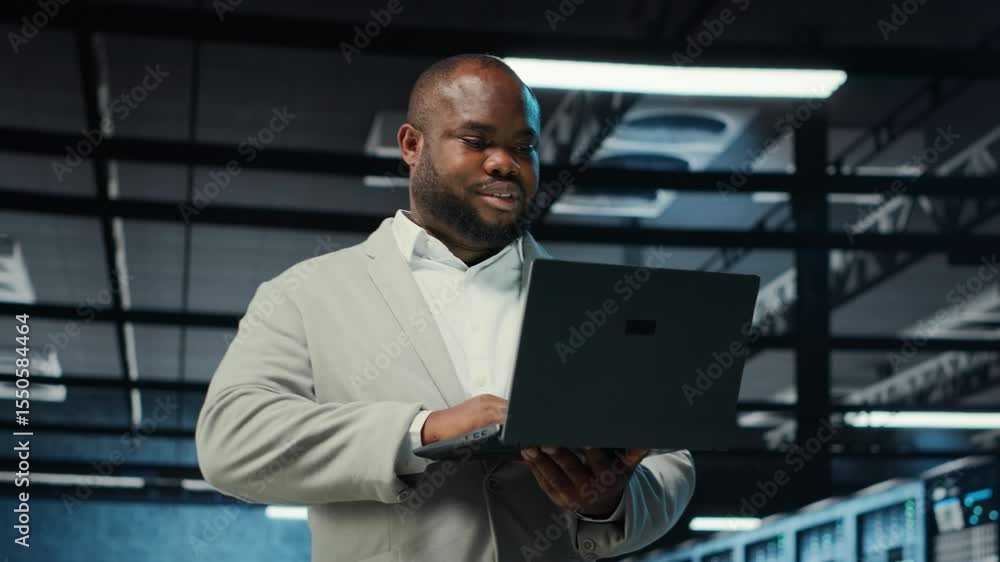 Poster engineer walking in server room backing up storage arrays using software on laptop. african american