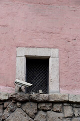 Surveillance Camera Overlooking a Shaded Window Set Against a Pink Wall with Stone Base: A Blend of Modern Security and Historical Architecture's Repose