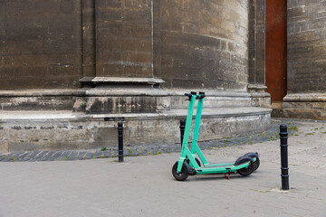 A Bright Green Electric Scooter Parked Beside Historic Stone Pillars on a City Sidewalk, Blending...