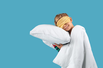 Cute little happy boy in pajamas with sleeping mask, pillow and blanket on blue background