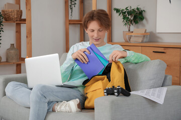 Male student with laptop putting copybooks into backpack on sofa at home
