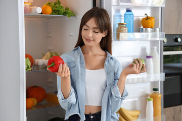 Young woman with bell pepper and apple near open fridge in kitchen