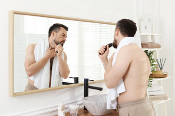Handsome man shaving with trimmer near mirror in bathroom
