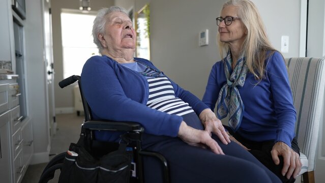 A woman in her 90s sits in a wheelchair, smiling as her daughter provides care and companionship during a visit at their cozy home together.