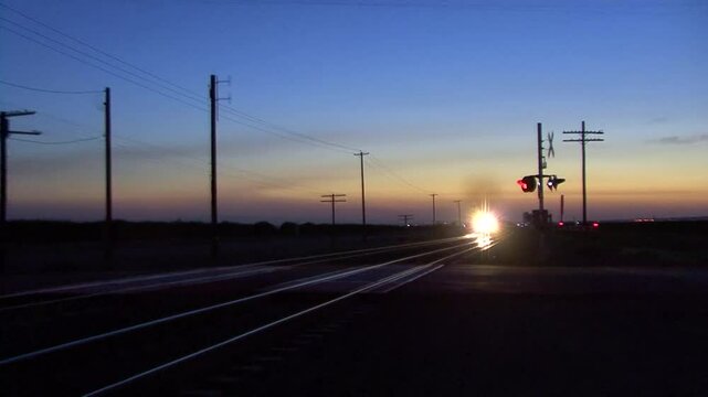 It's nightfall and a freight train approaches a railway crossing, lights blazing, horns blaring and wheels screeching while the crossing signals flash and ring as the train passes almost too close.