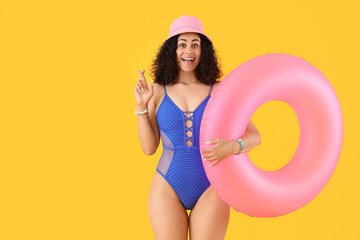 Young African-American woman in swimsuit with pink inflatable ring crossing fingers on yellow...