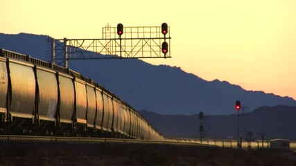 A long line of uniform railroad hopper cars file past, glowing in the last rays of the setting desert sun, against a silhouetted backdrop of layered mountains in the California Mojave Desert.