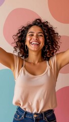 A joyful young woman with curly hair smiles brightly, arms raised, wearing hoop earrings and a beige tank top against a colorful, abstract background.