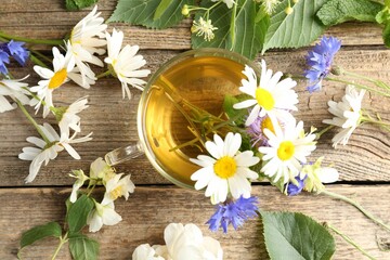 Aromatic herbal tea in glass cup, flowers and green leaves on wooden table, flat lay