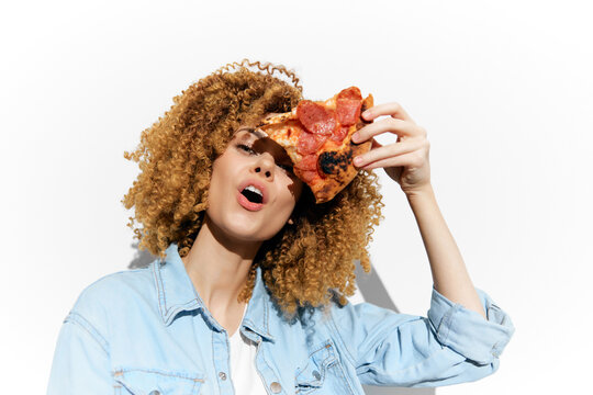 Cheerful young woman holding a pizza slice with pepperoni, expressing joy against a bright white background, ideal for food lovers and healthy eating concepts