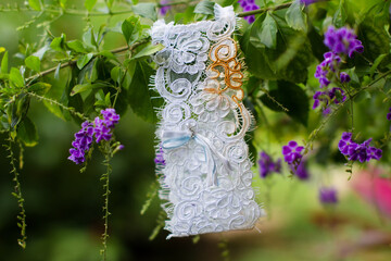 Lace bridal garter with blue ribbon detail hanging on a green branch with purple flowers. A...
