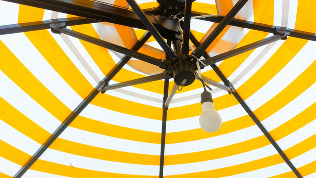 Looking up at a yellow-and-white striped patio umbrella; metal spokes radiate around a single hanging bulb, forming a striking geometric pattern against the bright canopy. - Powered by Adobe