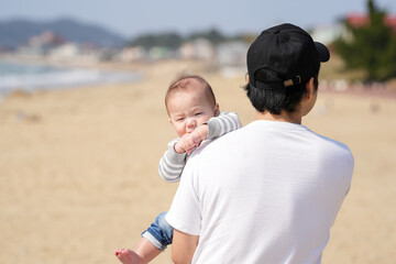 In early March, a Japanese father in his 30s carries his 6-month-old baby on his shoulders while walking along a sandy beach by the sea. A warm springtime scene of family, joy, and connection.