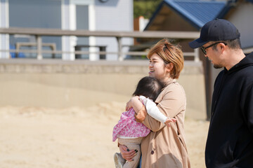 In early March, a 1-year-old Japanese girl and her parents in their 30s walk together on the sand in front of a beachside house. A cheerful springtime family moment with laughter and sea breeze.