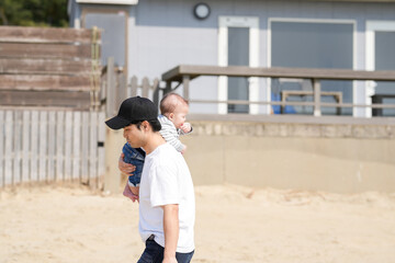 In early March, a Japanese father in his 30s carries his 6-month-old baby on his shoulders while walking along a sandy beach by the sea. A warm springtime scene of family, joy, and connection.