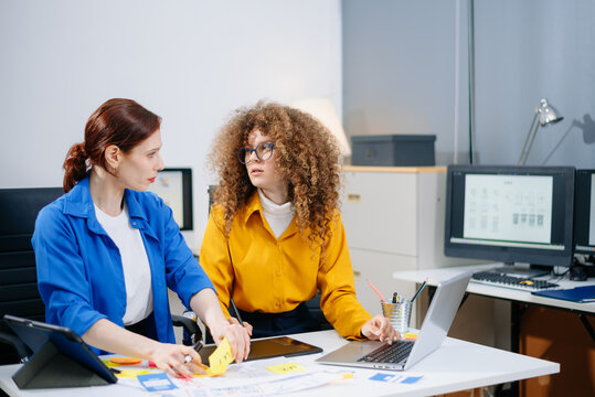 Two women discussing mobile app wireframes in a modern office. UX UI team collaboration, design thinking, and creative planning.