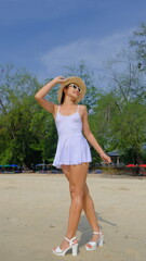 Stunning Asian woman in a short white dress and straw hat strolls confidently along a sunny beach