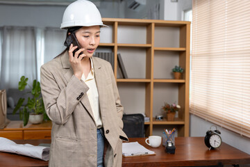 Young Asian female engineer wearing white safety helmet standing office talking mobile phone with...