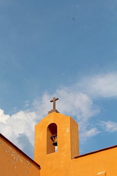 Peque&ntilde;o campanario sobre barda amarilla con remate de cruz y cielo con nubes.