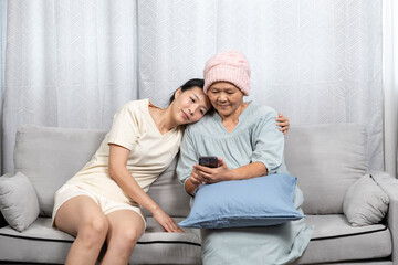Young Asian woman leaning head gently on middle-aged Asian female cancer patient wearing pink knitted hat using smartphone on sofa, showing peaceful companionship, love, digital connection, and care.