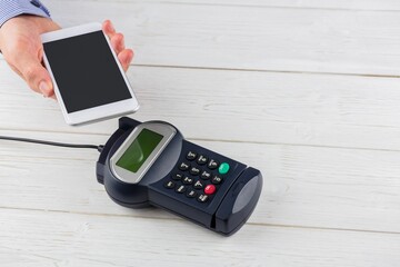 Man is holding smartphone above POS terminal with cable on pale wooden tabletop in flat design