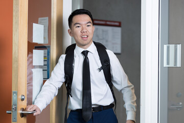 A young man in a dress shirt and tie enters through a door with a backpack. Suitable for intern,...