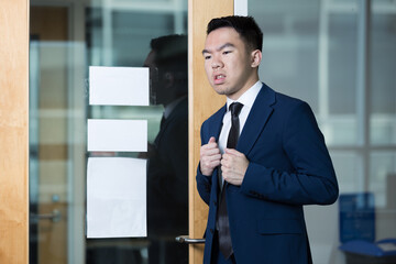 A young man in a suit stands outside a glass door, adjusting his jacket with a tense expression....