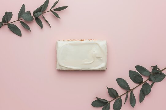 A single slice of white frosted cake is artfully placed on a pastel pink background with dark green eucalyptus branches on either side - Powered by Adobe