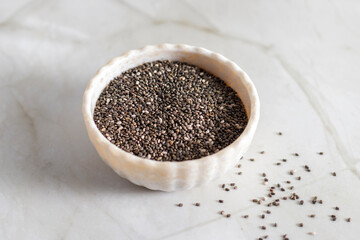 A white ceramic bowl filled with black chia seeds sits on a light marble surface, with some seeds scattered casually around the bowl’s base.
