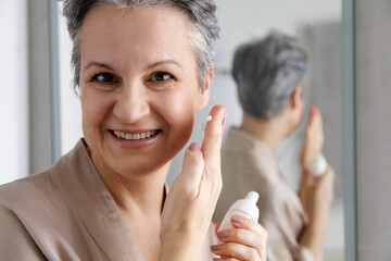Smiling senior woman applying face cream during daily skincare routine in front of mirror