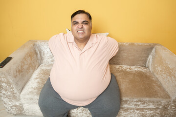 Overweight indian man suffer from backache sitting on couch. Plus size guy have pain in spine due to excess belly fat. Healthcare and Obesity problem. Top angle.