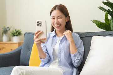 Happy brunette beautiful asian young woman wear casual sitting relax on couch, use mobile smart...