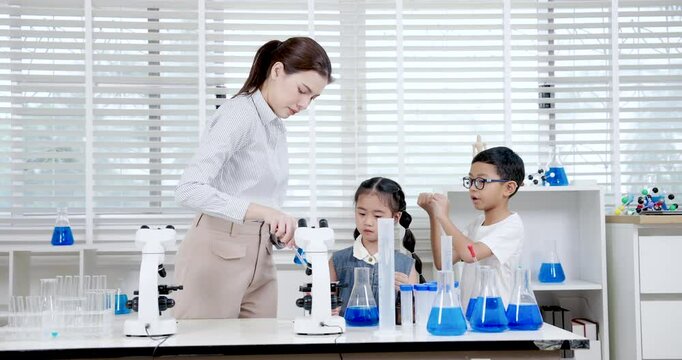 Asian female teacher assisting young girl with science equipment while young boy holding cylinder in primary science class during practical chemistry experiment using beakers and microscope