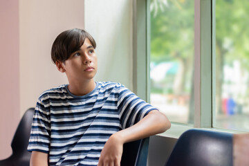 A young boy in a striped shirt gazing out the window with a curious and thoughtful look, seated indoors. Moment of imagination, dreaming, or waiting.