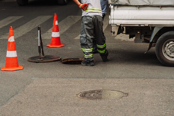 Urban utility workers servicing underground water infrastructure at an open manhole. Safety cones, service van, and tools visible. City maintenance without visible faces or brands