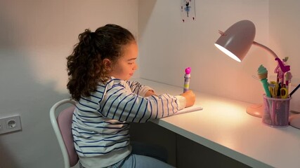 Little girl doing schoolwork in her room under the glow of a desk lamp, enjoying a creative moment while writing alone at night in a calm and well-organized home study space