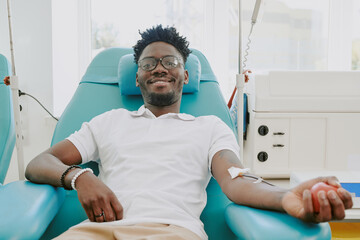 Young adult Black man donating blood while reclining in medical chair, smiling and looking relaxed,...