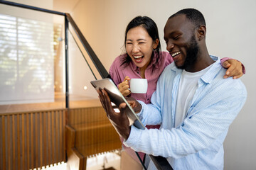 Multinational couple enjoying their morning routine, smiling and using laptop and tablet at home