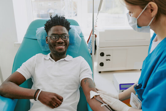 Black young adult man donating blood while sitting in medical chair, smiling at nurse preparing his arm with cotton swab, healthcare professional wearing mask and gloves - Powered by Adobe
