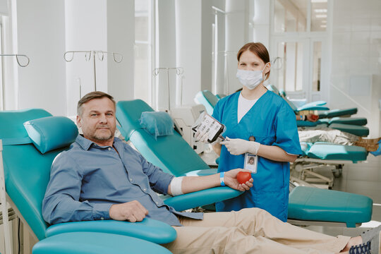 Middle aged Caucasian man donating blood while sitting in medical chair, holding red stress ball, young Caucasian female nurse wearing mask standing beside holding blood collection supplies