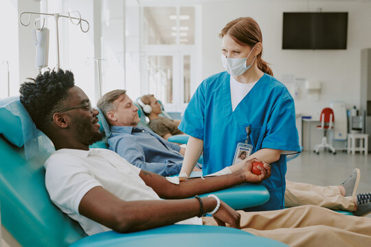 Young Black man donating blood while medical worker assisting, middle aged Caucasian man and young Caucasian man donating blood in background, hospital blood donation center setting