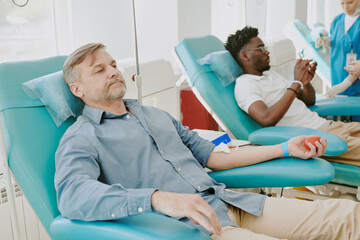 Obraz premium Middle aged Caucasian man and young Black man donating blood while reclining in medical chairs, both connected to intravenous lines, healthcare worker preparing equipment in background