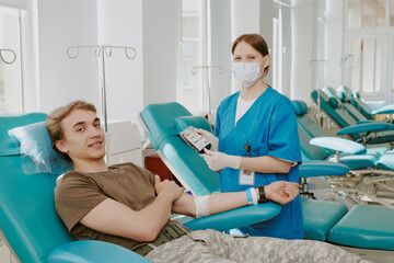 Obraz premium Caucasian young adult man donating blood while sitting in medical chair, female Caucasian nurse wearing mask and gloves holding blood bag, hospital setting with empty chairs visible